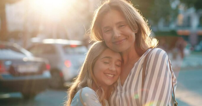 Portrait of happy mother and teen daughter standing hugging against the backdrop of street in sunlight and smiling at camera enjoying moment.