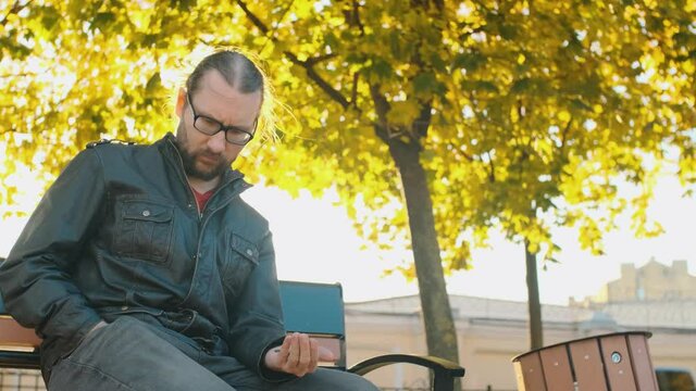 A poor man in old clothes and glasses counting coins in the palm of his hand sitting on the bench in the park. A broken person got into poverty, been laid off. Unemployment. Autumn trees background