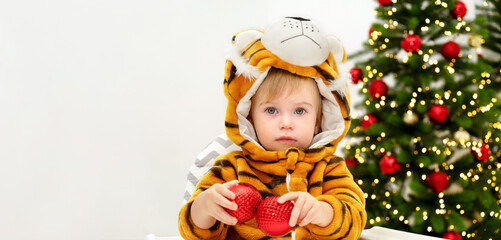 Cute baby wear costume of tiger,symbol of chinese New 2022 Year on background of Christmas tree sit on chair,play with balls,have fun.Infant celebrate holiday at home.Child Masquerade idea.Copy space