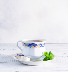 A porcelain teacup with a saucer stands on a light wooden table. Blue and gold pattern on the cup. Sugar cubes and mint leaves on a saucer
