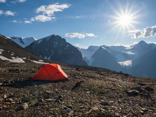 Tent on the edge of a cliff. Camping on a rocky high-altitude plateau. Orange tent on the background of high snow-capped mountains.