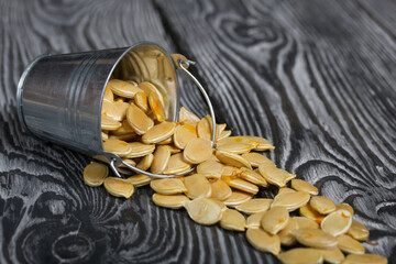 A metal bucket filled with pumpkin seeds. The bucket is overturned, the seeds are scattered over the surface. On black pine boards. Close-up shot.