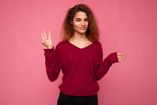 Photo of young positive attractive brunette wavy lady with sincere emotions wearing casual pink sweater isolated over pink background with copy space and showing three fingers