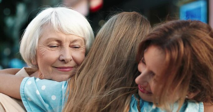 Close up of happy caucasian teenage girl standing on street background and tenderly hugging her mother and grandmother in farewell.