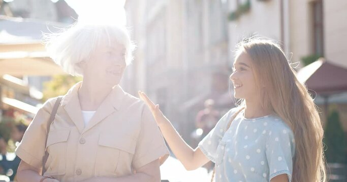 Beautiful gray-haired woman stands on street and looks at clock waiting for meeting with her cute granddaughter.