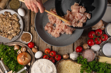 The cook is pan frying pieces of chicken fillet. Ingredients. Lots of facilities. Beautiful composition. The look of a high angle. Cooking pizza, pie, salad, ravioli. Wooden texture.