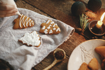 Hands decorating gingerbread cookie christmas tree with frosting on rustic table with napkin, candle, spices, decorations. Atmospheric moody image. Making traditional christmas gingerbread cookies
