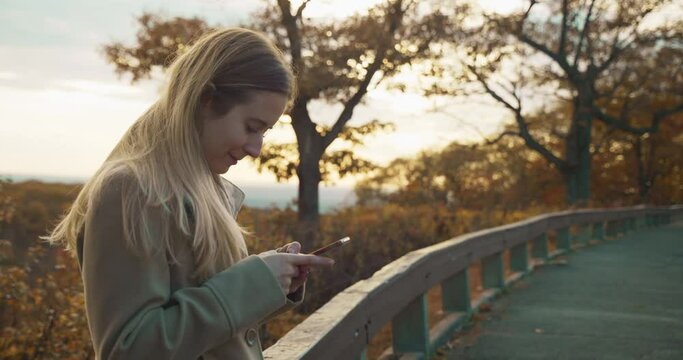 Young Woman Texting Typing Emailing Social Media on Cell Phone Smartphone and Smiles with Fall Autumn Trees and Leaves on top of Mountain