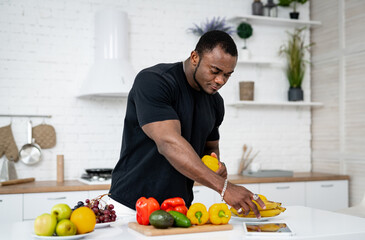 Multiracial man standing at the kitchen full of fruits and vegetables in the modern interior and looking at the tablet. Food and health care concept