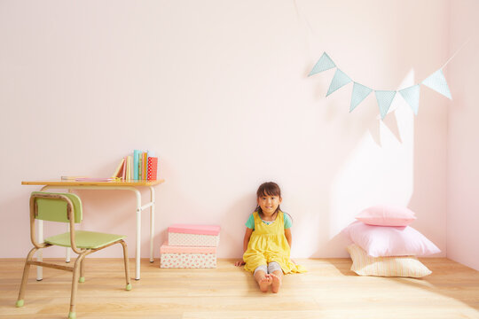 ピンク色の壁の子供部屋で勉強机の横の床に座っている女の子