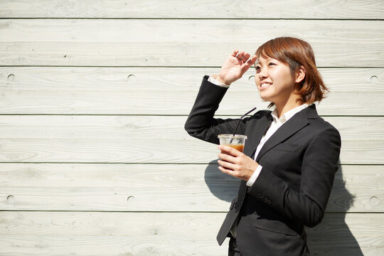 コーヒーを片手に日差しをもう片方の手で避けるスーツ姿の女性