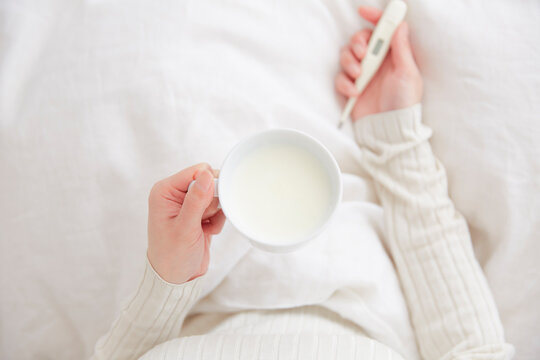 ベッドの上でホットミルクのカップと体温計を持つ女性の手元