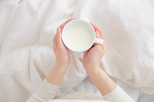ベッドの上でホットミルクのカップを持つ女性の手元