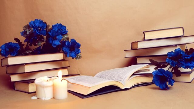 Old books and blue flowers isolated on beige craft background. An unfolded book near a candle with a restless flame. Literary background.
