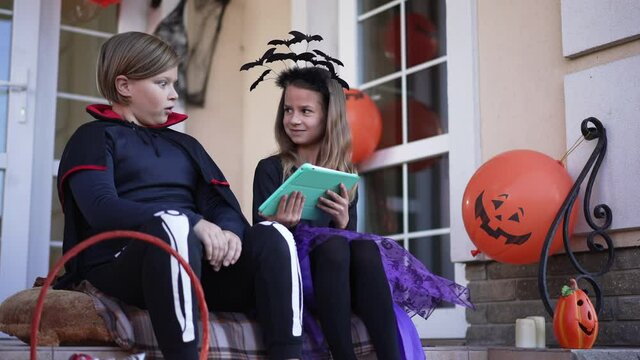 Scared boy with frightened facial expression looking at girl showing horror on tablet smiling. Positive Caucasian children in costumes having fun in slow motion sitting on porch on Halloween