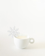 White snowflake on white coffee cup with milk or hot drink against white background. Winter holiday layout. New Year or Christmas aesthetics concept.