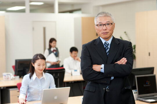 オフィスの中に立って腕組みするスーツを着た中高年社長