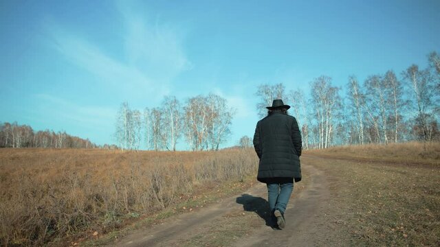 On a sunny autumn day, a lonely city man walks along a rural road. He is wearing a hat and a dark jacket. He has nothing with him. No one knows where he is heading his way. Beautiful autumn landscape.