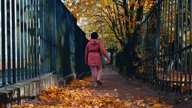 Young Caucasian Girl Walking Alone Narrow Paved Alley Between Metal Fences Kicking Heap of Yellow Withered Autumn Leaves on Sunny Windy Day