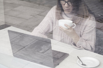 Young attractive woman working on laptop in modern coworking space and drinking cocoa. Elerning or freelance concept. View through the glass window