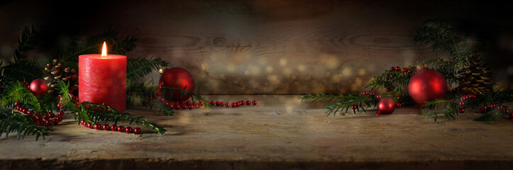 Atmospheric Advent and Christmas decoration with a lit red candle, balls and evergreen yew branches on dark rustic wooden planks, wide panoramic format, copy space