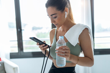 Fitness woman using mobile phone and drinking water while training on exercise bike at home.