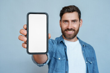 Fototapeta na wymiar Satisfied confident smiling adult businessman showing mobile phone blank white screen mockup to camera standing isolated on blue background. Man holding smartphone and shows copy space for advertising