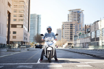 Wide shot of a young motorcyclist stopped at a traffic light in Barcelona. The man riding his scooter through the city on a large avenue lined with skyscrapers - obrazy, fototapety, plakaty
