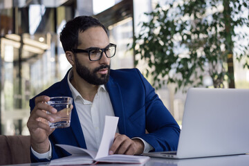 Handsome confident arabic businessman planning start up project, holding glass of water in modern...