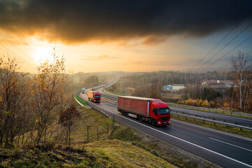 Red trucks driving on the highway winding through forested landscape in autumn colors at sunset