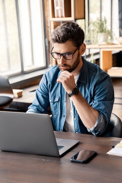 Handsome Focused Man Working Using Laptop Sitting At Office Desk Table In Coworking Workspace. Businessman Freelancer, Executive Manager Or Employee Making Report, Searching Information. Vertical Shot