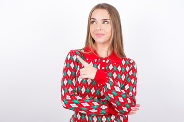 Portrait of Young beautiful caucasian girl wearing christmas sweaters on white background posing on camera with tricky look, presenting product with index finger. Advertisement concept.