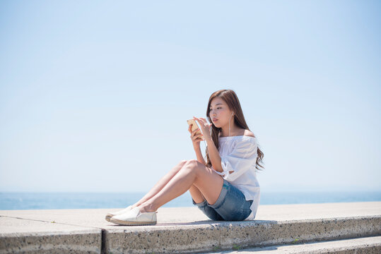 海の堤防に座ってスマホで音楽を聴く女性