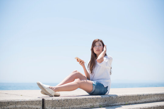 海の堤防に座ってスマホで音楽を聴く女性