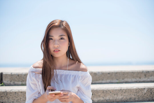 海の堤防に座ってスマホで音楽を聴く女性