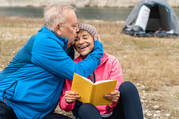 Happy senior couple having tender moment reading a book while camping in the woods - Elderly lifestyle - Focus on woman face