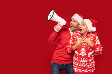 Happy beautiful mature couple in christmas sweaters and santa claus hats shouting on megaphone.