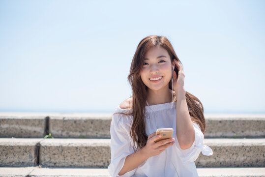 海の堤防に座ってスマホで音楽を聴く女性