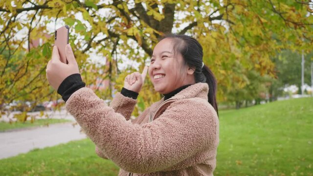 Happy Asian woman taking video call on smartphone at the park to show the view in Autumn, woman wearing light brown coat, yellow leaf on the tree, Beautiful day in Autumn season, Sweden