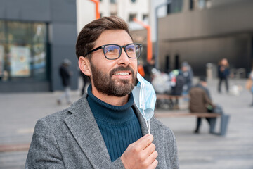 Happy relaxed businessman taking off protective medical mask from face outdoors