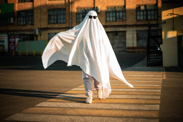 Celebrating halloween. Ghost Challenge 2021. A man dressed as a ghost from a sheet and sunglasses crosses the road along the crosswalk. Spooky season.  - obrazy, fototapety, plakaty