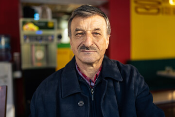Front view portrait of senior caucasian man with mustaches sitting in cafe alone looking to the camera wearing coat close up copy space