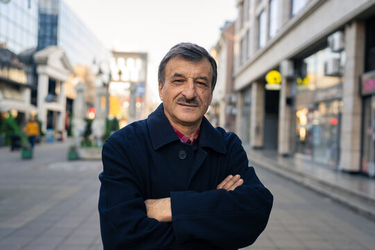 Front view portrait of senior caucasian man with mustaches standing in the city in winter or autumn day looking to the camera wearing coat close up copy space
