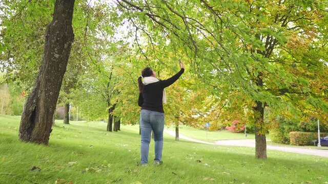 Asian woman wearing black long-sleeved shirt and scarf, trying to take a picture of tree at park in Autumn , using smartphone taking a photo, Beautiful day in Autumn season, sweden