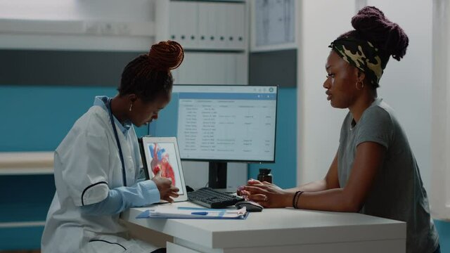 Doctor pointing at tablet with cardiovascular figure, explaining heart condition and blood flow to patient. Medic showing device screen with cardiac organ and cardiology diagnosis.