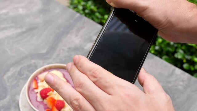 Man, influencer, blogger shoot on phone camera breakfast, restaurant food. Male hands making photo, picture, shots for social media of healthy fruit and berries smoothie bowl on smartphone. Close-up.