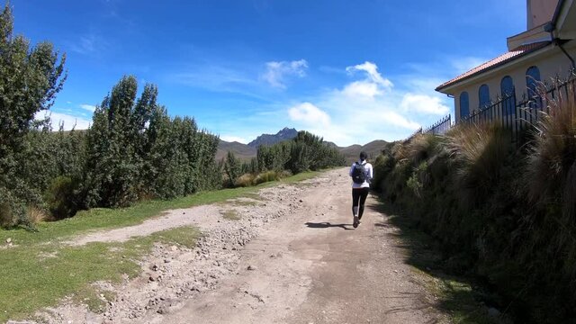 A woman with a black backpack is walking on a trail of dirt next to polylepis trees and a church on the moorlands of the Andes in Rucu Pichincha, Ecuador.