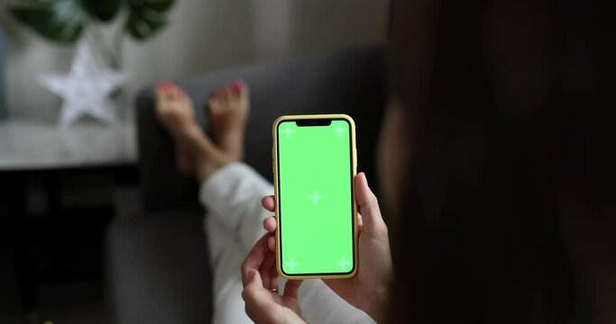 Young woman using mobile phone with green screen for copy space closeup. Chroma key mockup on smartphone in hand. Person holds cellphone and swipes photos or pictures indoors at home