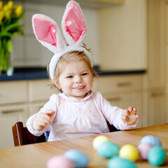 Cute little toddler girl wearing Easter bunny ears playing with colored pastel eggs. Happy baby child unpacking gifts. Adorable healthy smiling kid in pink clothes enjoying family holiday