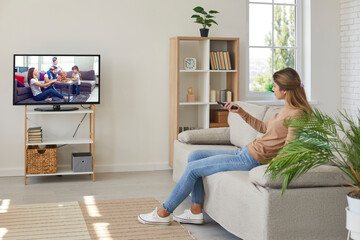 Young woman renter sit rest on couch in living room browse TV program with remote control....